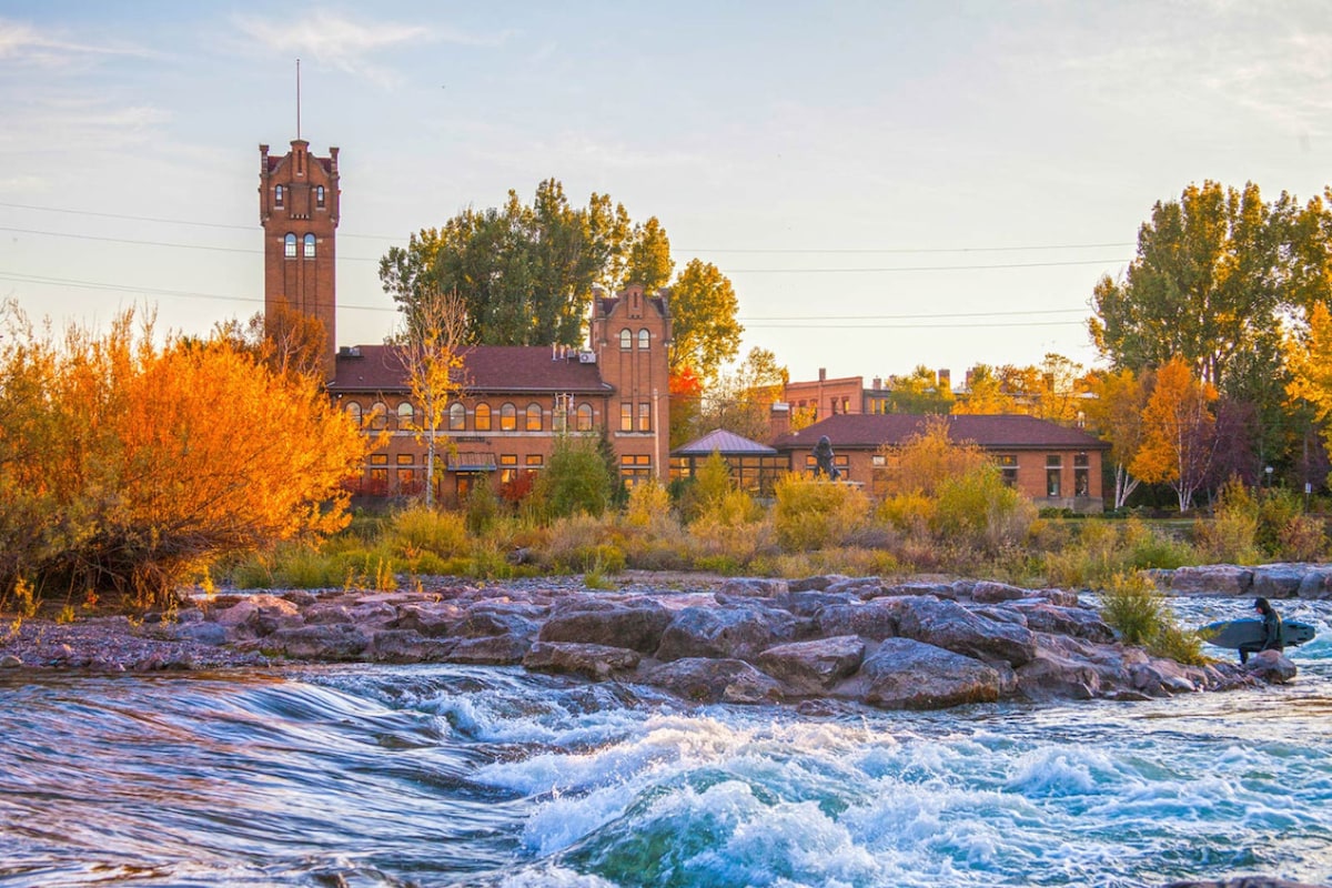 A historic building with a distinctive tower is positioned beside a flowing river. Vibrant foliage in autumn shades surrounds the area, with smooth rocks partially submerged in the water. The scene is framed by a clear sky as the day transitions to evening.