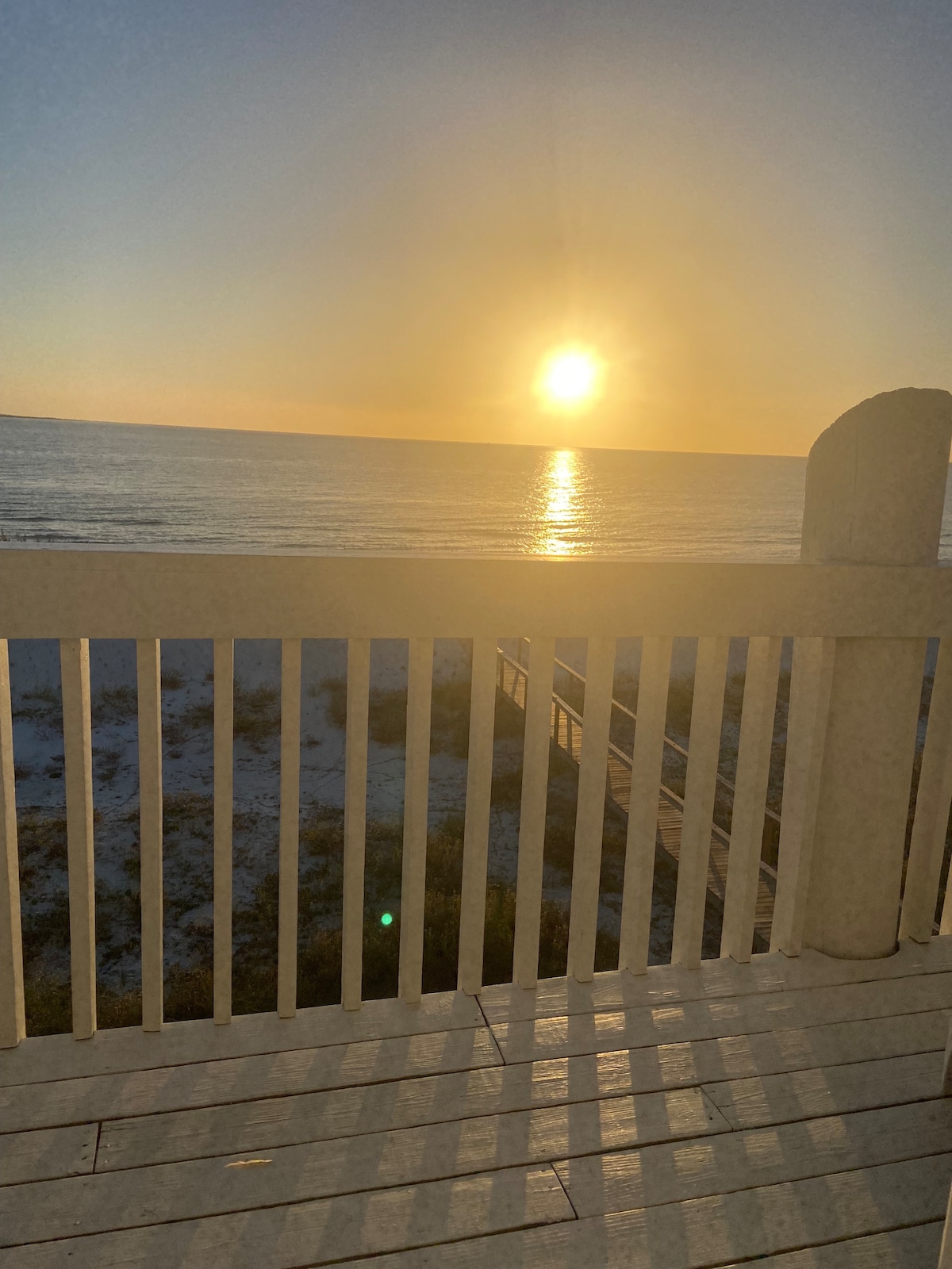 A tranquil sunset is captured over the ocean, with the sun reflecting on the water's surface. The image includes a portion of the balcony railing, framing the serene view of the shoreline and the soft, warm colors of the evening sky.