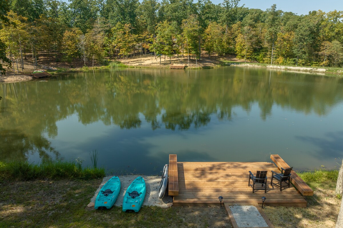 The lakefront area features two kayaks positioned beside a wooden dock and seating arrangement. The calm water reflects the surrounding trees, creating a serene atmosphere. Natural pathways lead away from the dock into the wooded landscape, providing easy access to the outdoors.