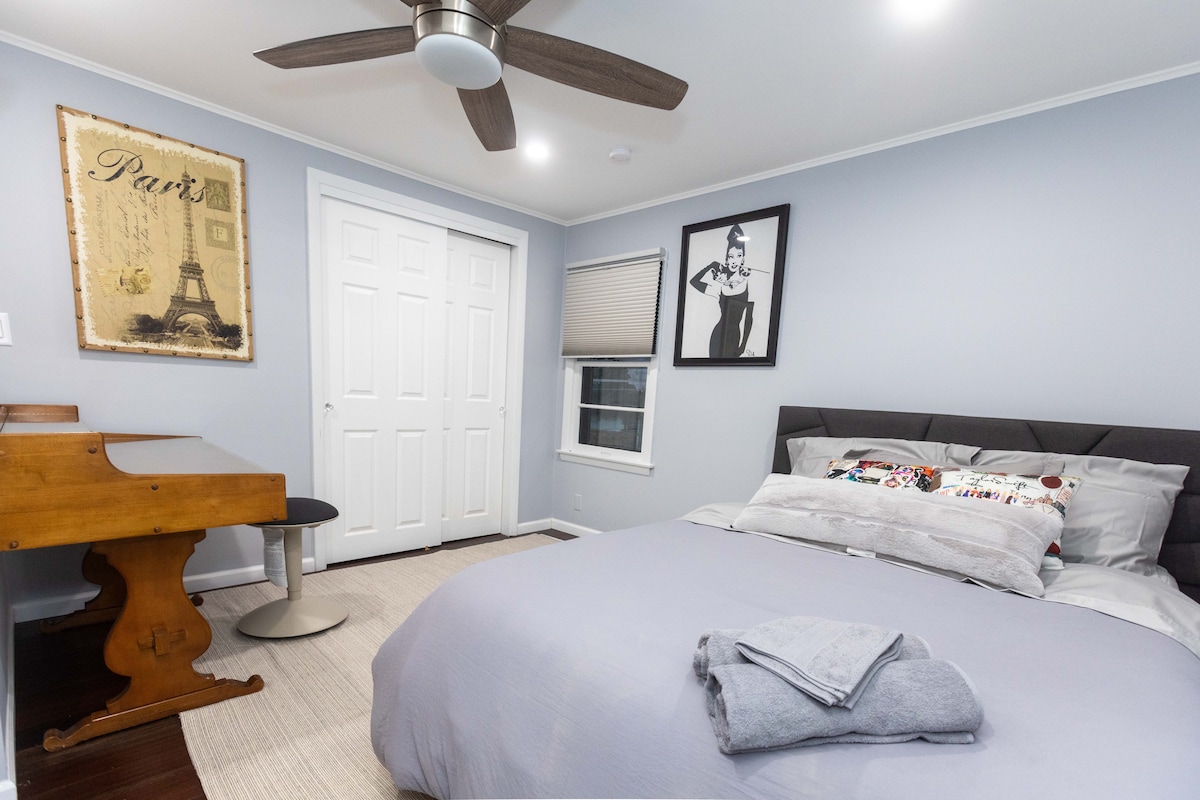 A private bedroom features a spacious bed with soft linens and decorative pillows. A work desk and modern chair are positioned nearby. The room is illuminated by a ceiling fan, and two closets provide ample storage, with light gray walls enhancing the overall calm atmosphere.