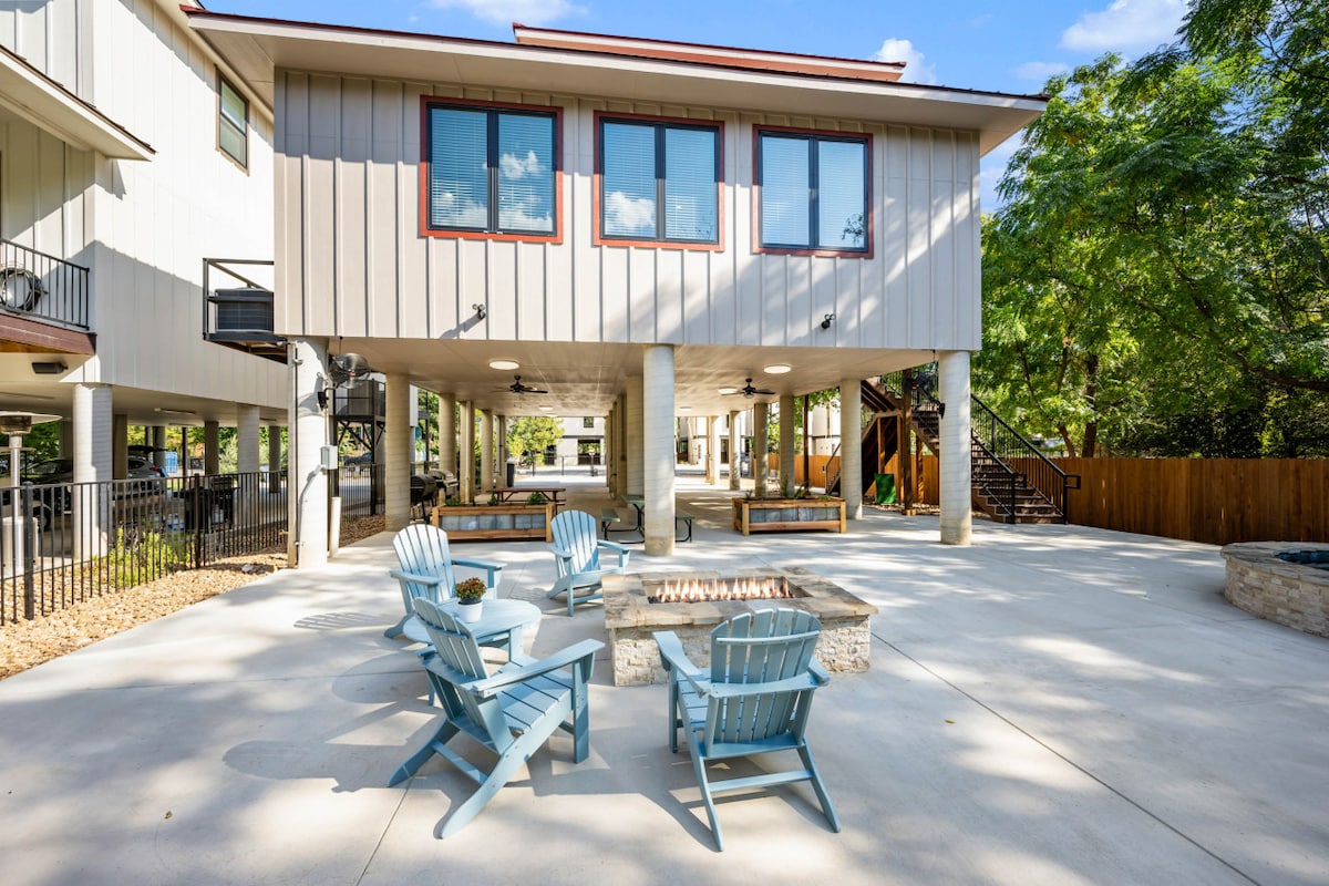 The outdoor living area features a spacious patio, highlighted by a large stone fire pit surrounded by several light blue Adirondack chairs. The elevated house provides a shaded area beneath, with a blend of concrete and natural landscaping that showcases trees and access to additional outdoor space.