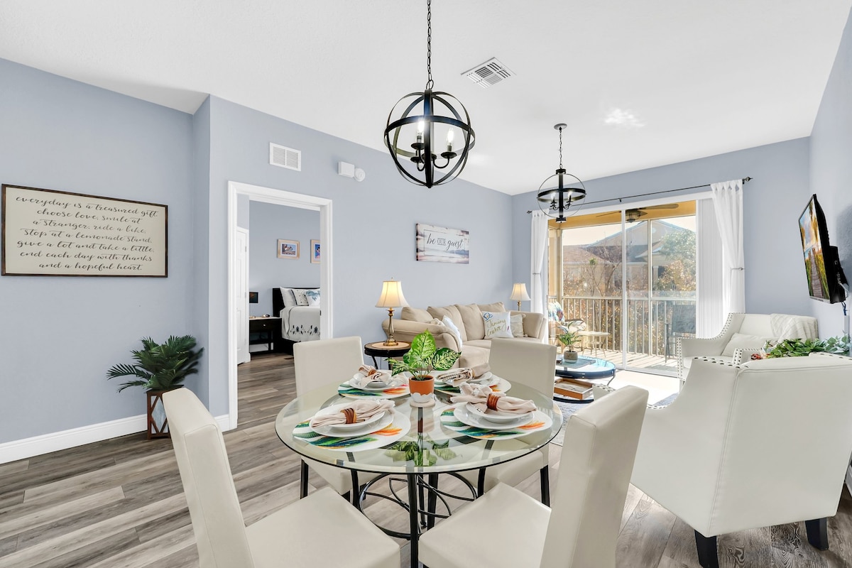 An open-concept living area is presented, featuring a circular dining table set for four, with colorful dishes and glasses. A cozy couch is positioned near large glass doors, leading to a balcony. Soft pastel walls complement modern light fixtures, creating a bright ambiance.