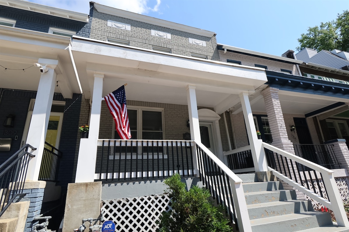 The exterior of the rowhouse is shown, featuring a covered porch with white columns and a welcoming front steps. An American flag is displayed, while well-maintained landscaping enhances the curb appeal. The overall design reflects a traditional neighborhood aesthetic.