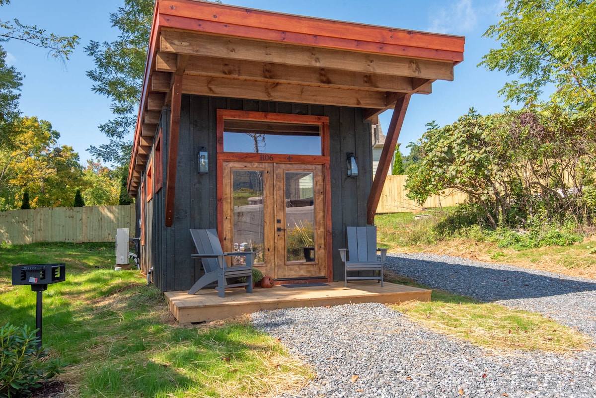 A modern cabin exterior features a wooden façade with a sloped roof. Two large doors are framed by decorative trim, flanked by adirondack chairs on a wooden porch. A gravel pathway leads to the entrance, surrounded by a neatly maintained lawn and greenery.