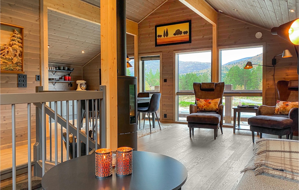 An inviting living area features large windows offering expansive views of the surrounding countryside. Two comfortable armchairs, adorned with decorative cushions, are positioned near a circular coffee table with candles. A dining table is visible in the background, enhancing the communal atmosphere.