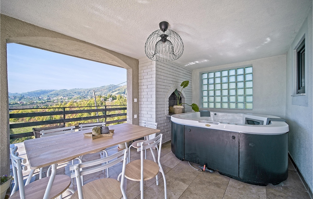 An outdoor terrace features a dining table with light wooden chairs, complemented by a jacuzzi tucked into the corner. Natural light filters through a large window, offering views of the surrounding hills and countryside.