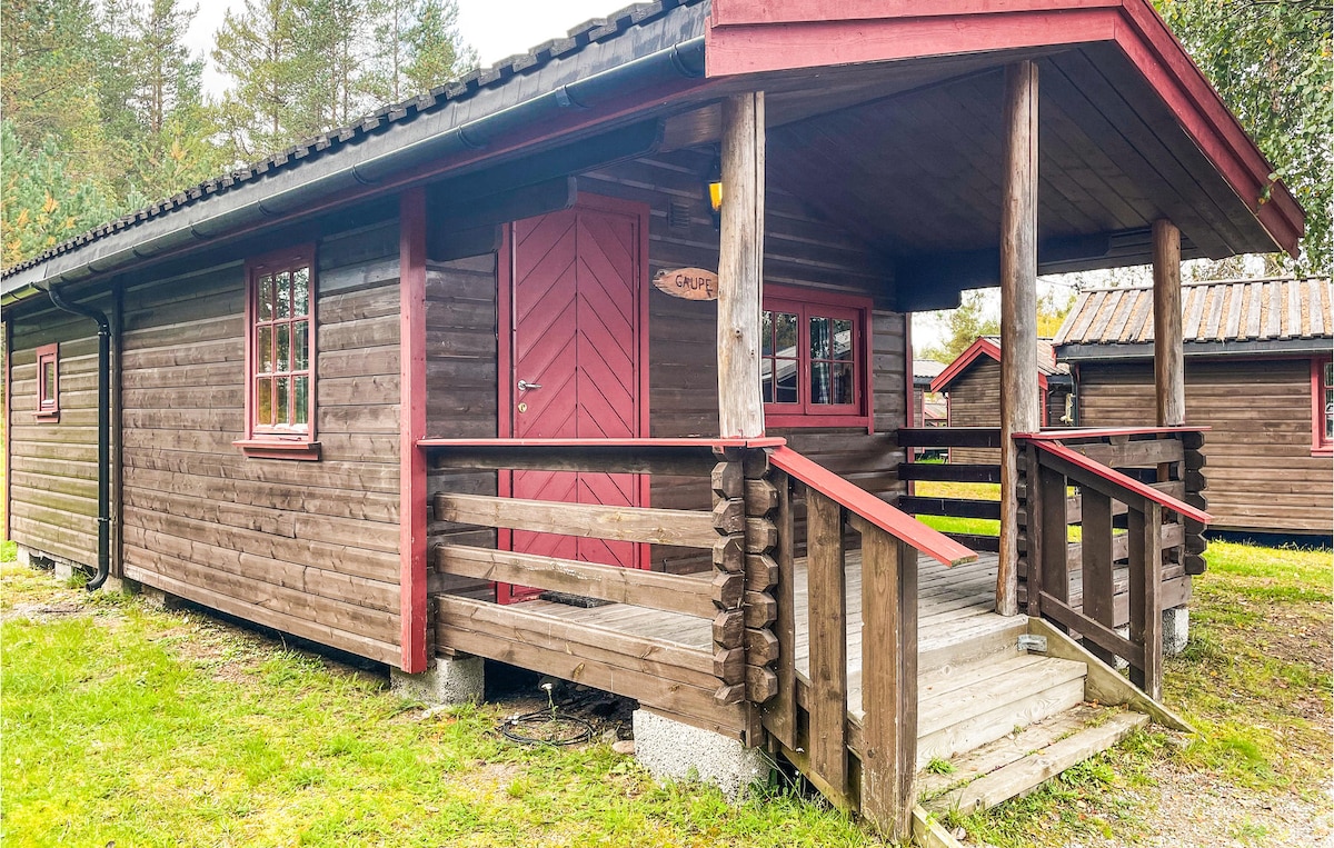 A wooden cabin with a rustic design features a red door and a covered porch. Railings encase the porch, and a path leads to the entrance. Surrounding greenery contributes to a tranquil outdoor setting.