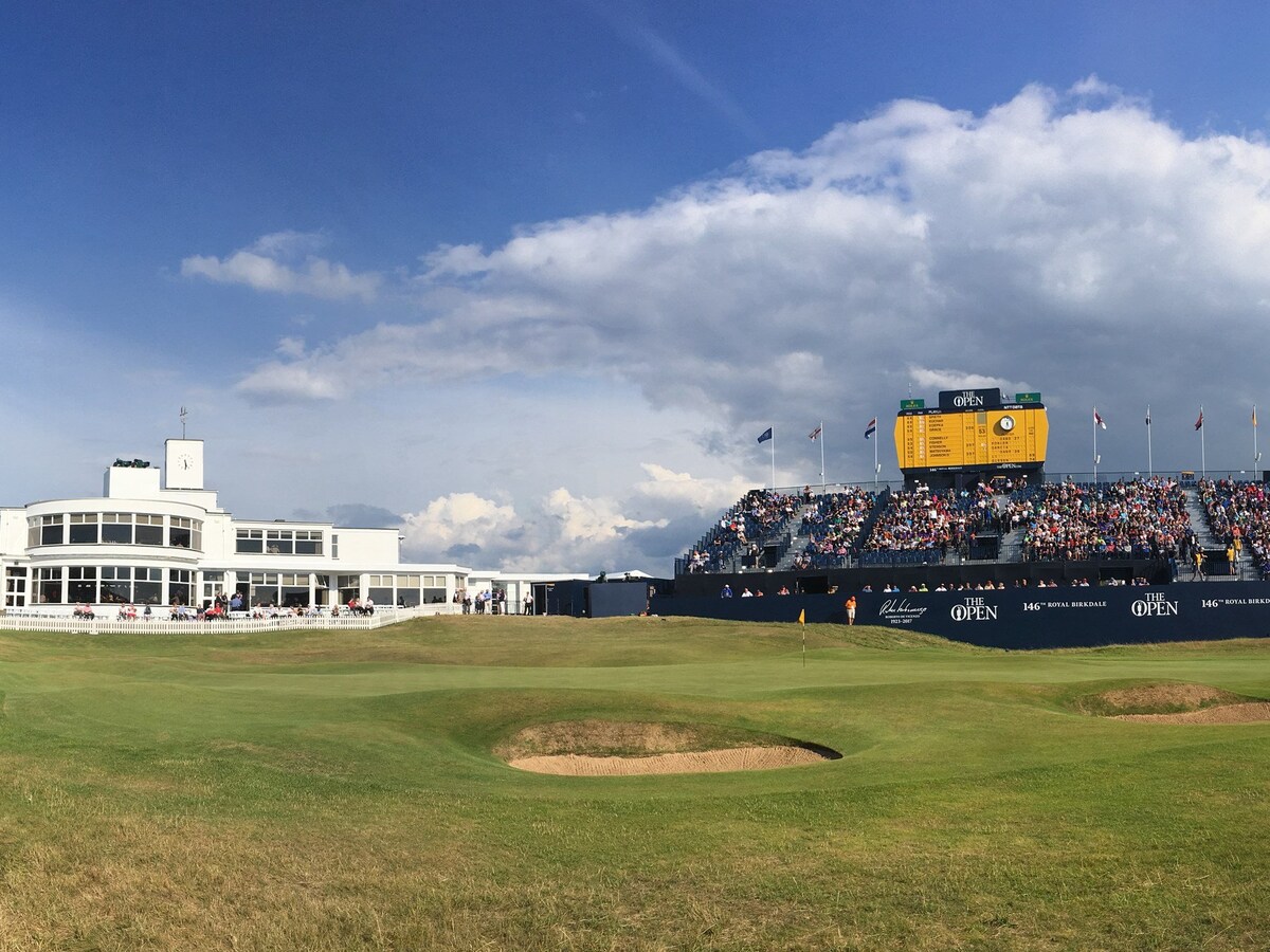 A spacious golf course is captured from a broad perspective, showcasing well-maintained greens and a prominent club building in the background. A large crowd can be seen in the grandstand, under a partly cloudy sky. The scoreboard displays multiple flags and event information.