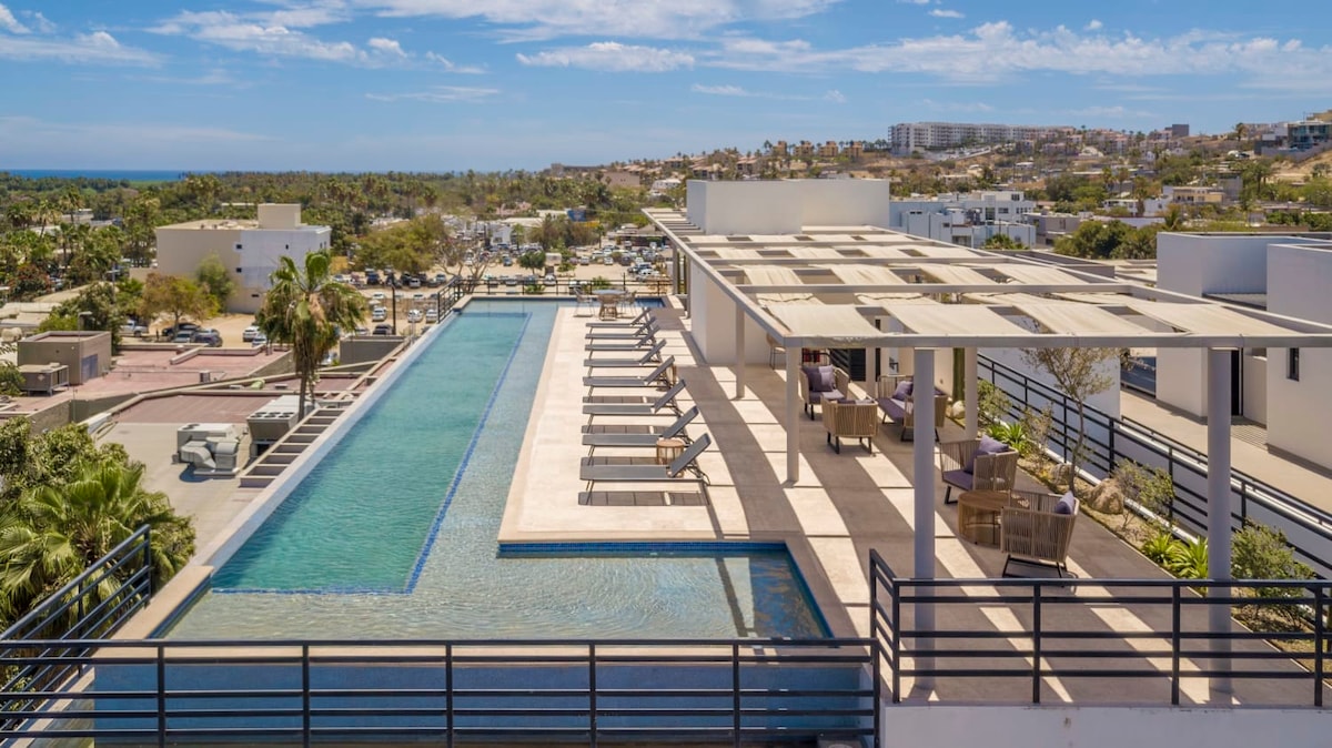 A spacious rooftop terrace features a long pool alongside comfortable lounge chairs. Sunlight reflects off the water, while shaded seating areas provide relaxation options. Scenic views of the surrounding area and the ocean are visible in the distance, enhancing the inviting outdoor space.