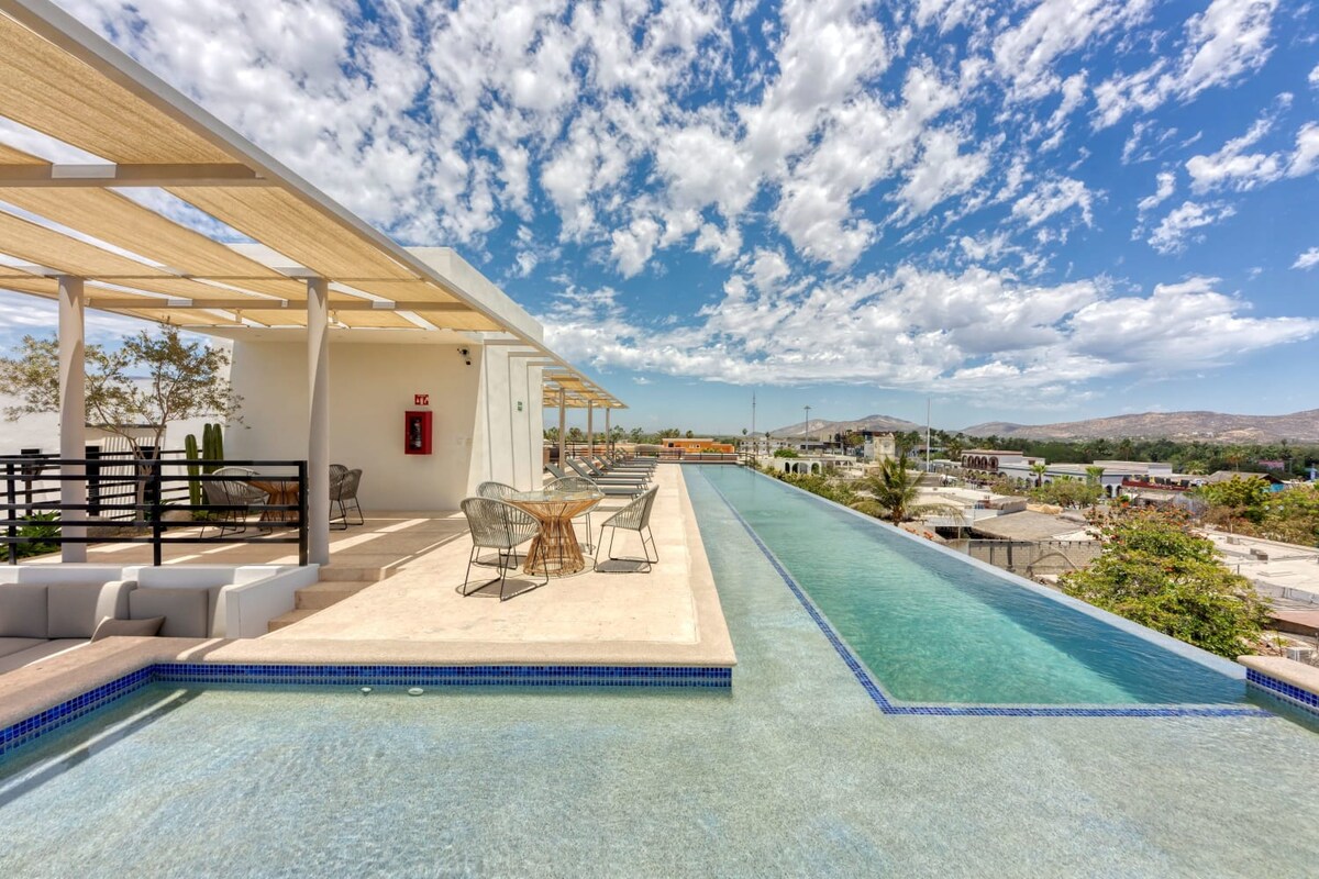 An exclusive rooftop terrace is shown featuring a long infinity pool that extends into the horizon, framed by a sleek lounge area with modern seating. Framed by clouds and blue sky, the landscape reveals a view of the surrounding area and distant mountains.