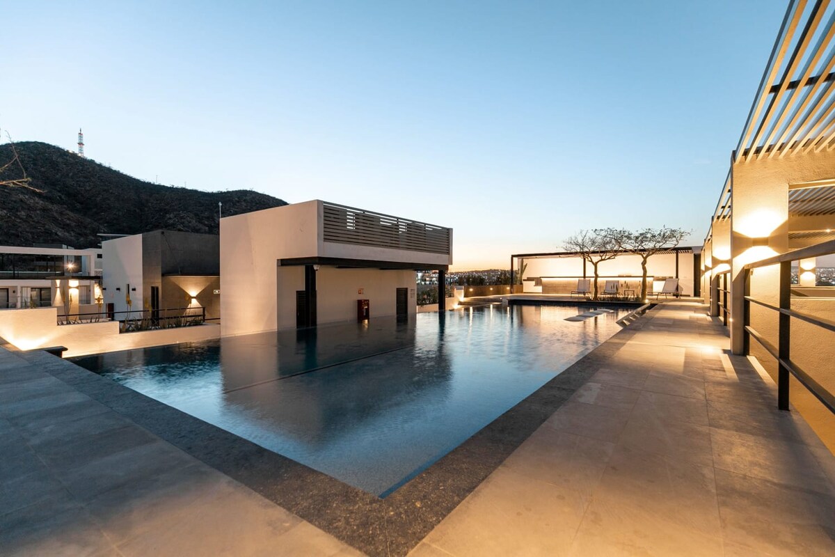 A spacious rooftop pool is surrounded by modern architecture, with gentle lighting reflecting off the water's surface. Comfortable lounging areas and a shaded seating space are visible nearby, set against a backdrop of distant hills under a clear evening sky.
