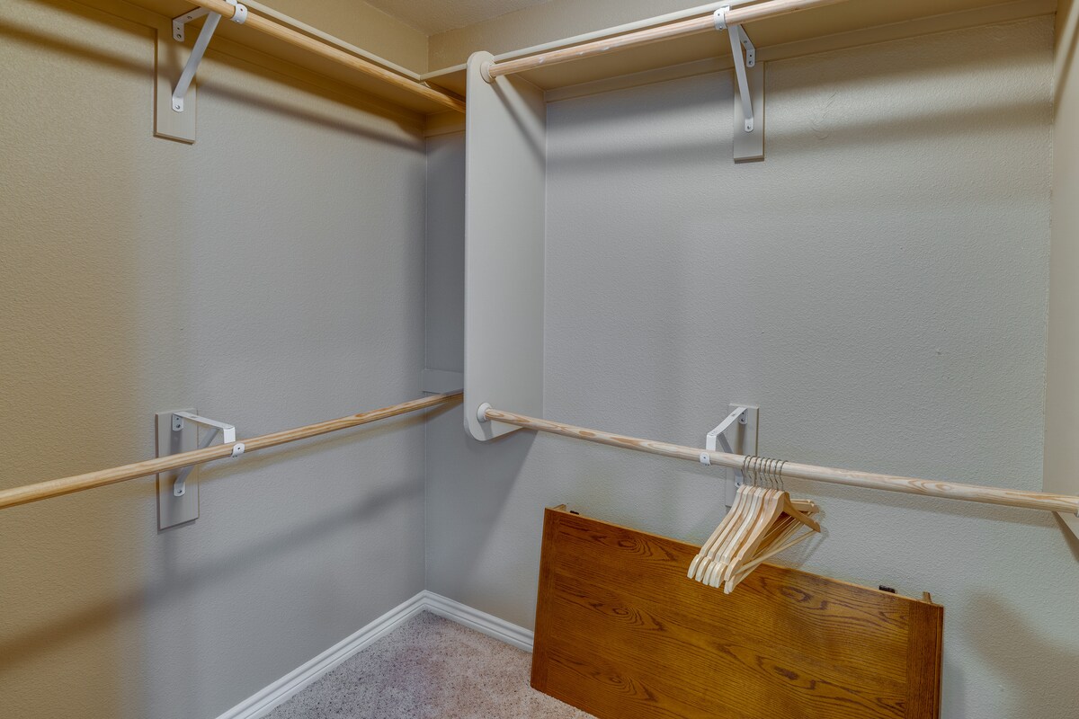 A spacious walk-in closet is presented with multiple hanging rods and a wooden shelf. Several wooden hangers are neatly arranged, while a wooden board leans against the wall, contributing to the organized appearance.
