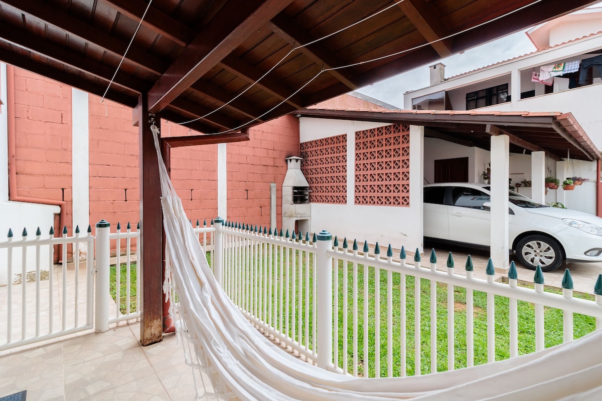 An outdoor area features a covered porch with a hammock, surrounded by a white picket fence. A green lawn is visible, along with a charcoal BBQ grill and a white car parked nearby. The backdrop includes a textured brick wall and a portion of the house.
