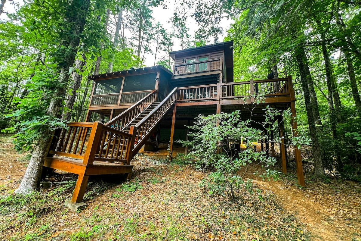 The cabin is nestled among lush greenery, featuring a rustic wooden exterior. A set of stairs leads to a spacious upper deck, while a screened-in porch can be seen on the main level. The surrounding trees provide a serene atmosphere.
