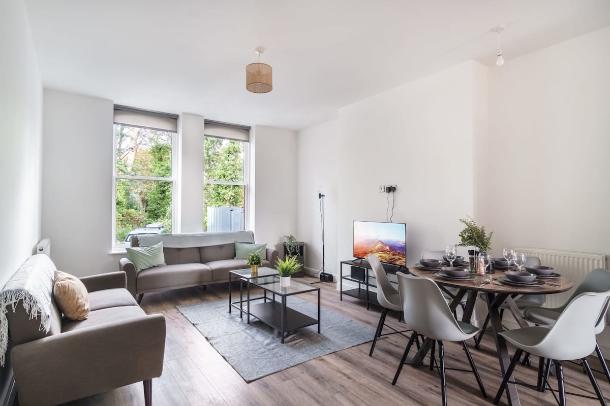 The open plan living area features two spacious sofas arranged around a coffee table, with a smart TV positioned nearby. A dining table is visible, set for multiple guests. Large windows allow natural light to fill the room, and greenery can be seen outside.
