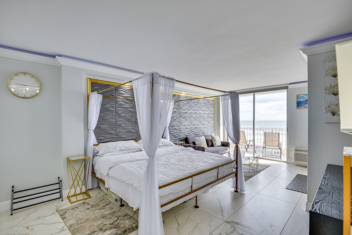 A spacious studio features a king bed with a canopy frame, complemented by a sleeper sofa. Large windows provide natural light and showcase views of the outdoor balcony. A round table and chairs are situated near the windows, enhancing the open layout.