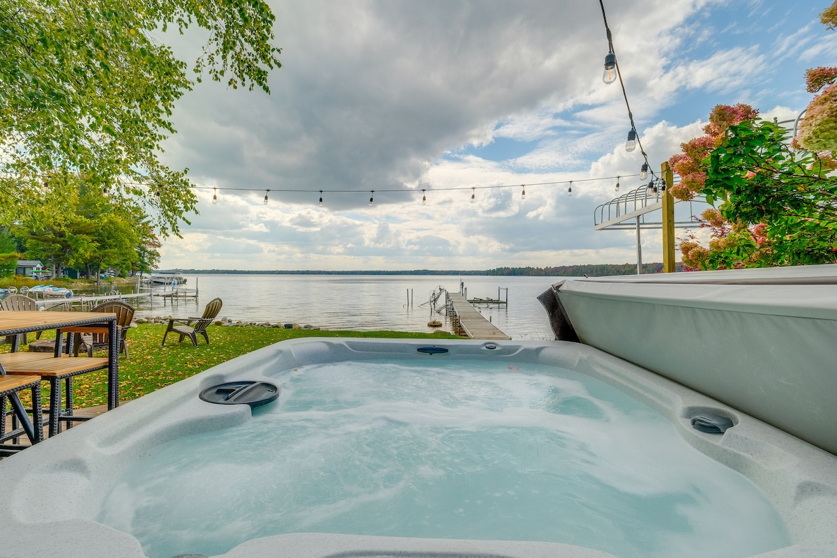 A private hot tub is positioned overlooking the lake, surrounded by greenery. Adorned with bistro lights overhead, the outdoor space offers a relaxing ambiance. A seasonal dock extends into the water, providing scenic views of the calm lake under a partially cloudy sky.