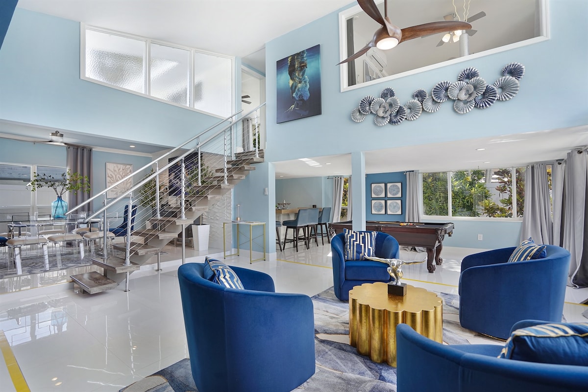 A spacious living area is depicted, featuring blue accent chairs arranged around a gold coffee table. The gleaming white floors reflect natural light from large windows. A staircase with a modern railing leads to an upper level. Artwork adorns the walls, enhancing the inviting atmosphere.
