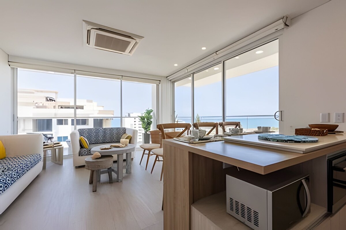 A bright living space is highlighted by large glass windows offering expansive ocean views. The room contains a comfortable sofa and stylish chairs surrounding a wooden coffee table. A modern kitchen is visible, featuring a countertop and appliances, enhancing the open layout.