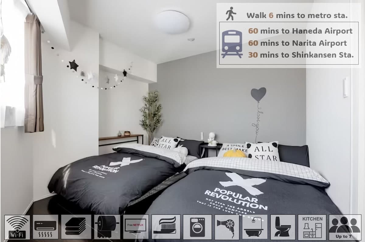 The spacious room features two individual semidouble beds with black bedding adorned with decorative pillows. A minimalist grey wall complements the light filtering through a window with sheer curtains, creating a calming sleeping area. A small plant adds a touch of greenery to the decor.