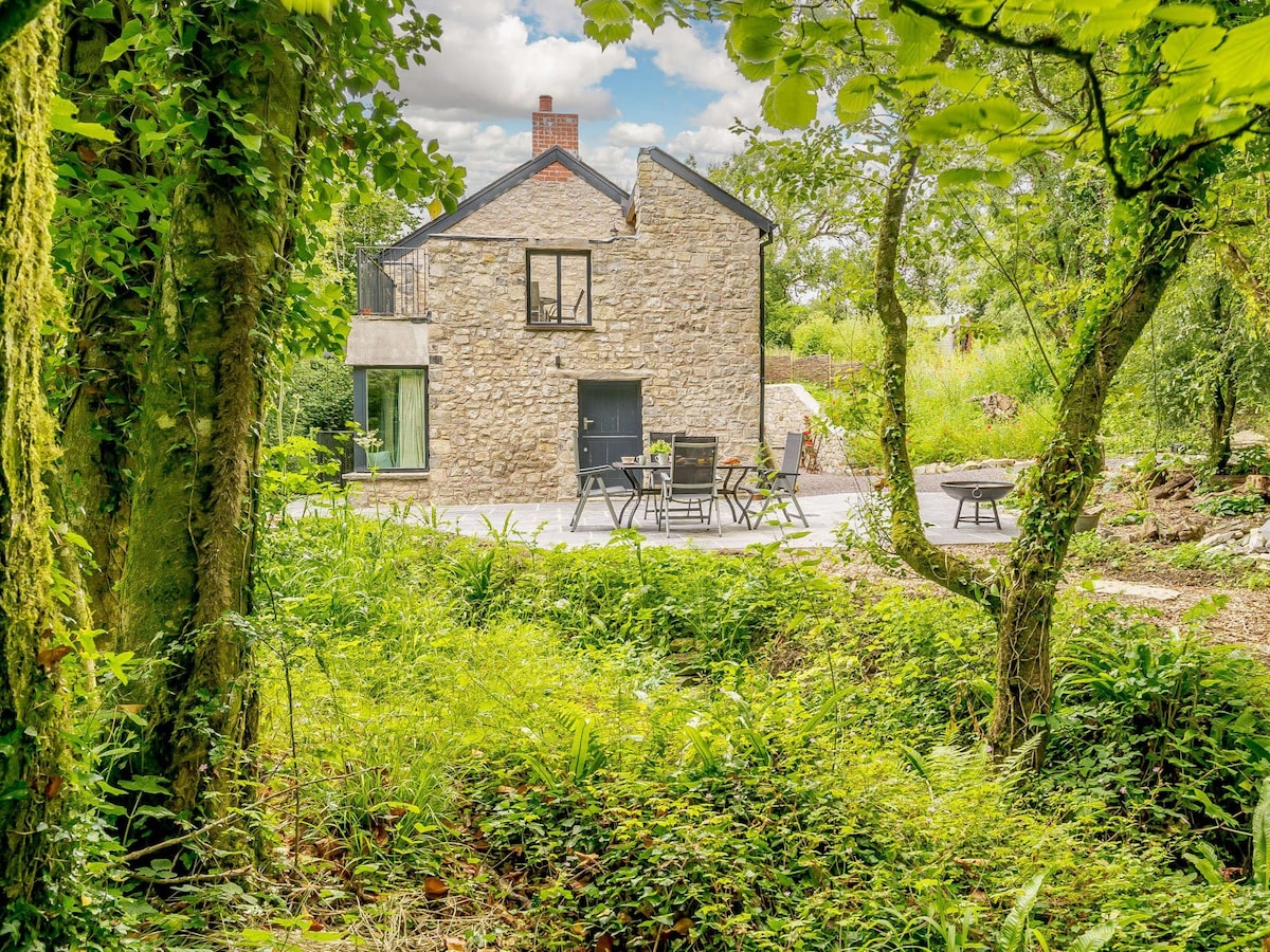 The exterior of a stone cottage is framed by lush greenery, with a spacious patio featuring a dining table and chairs. The property is set against a backdrop of trees and natural foliage, creating a serene, secluded atmosphere.