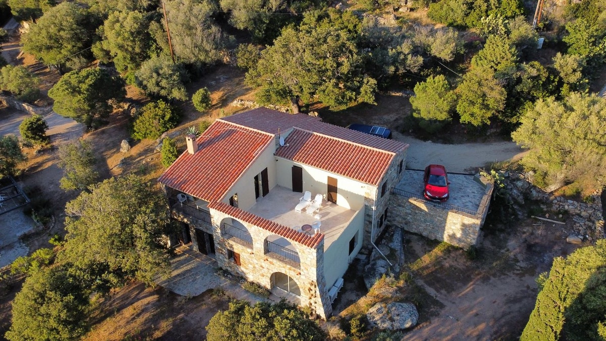 An aerial view showcases a charming apartment situated amidst lush greenery. The structure features a rustic stone exterior with a terracotta tile roof. A private terrace is visible, along with a modern car parked nearby, enhancing the sense of rural tranquility.