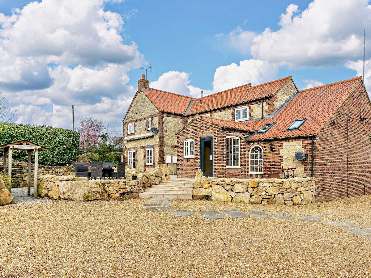 A charming country home is depicted, featuring a combination of stone and brick walls with a red-tiled roof. A gravel pathway leads to the entrance, flanked by a landscaped garden and seating area, inviting relaxation in a serene outdoor setting.