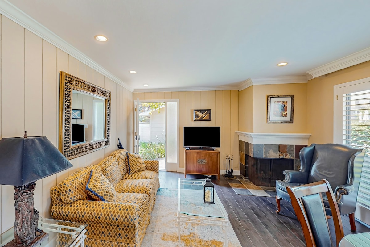 A bright and welcoming living area features a patterned sofa and a matching armchair. Natural light streams in from the patio door, illuminating the space. A smart TV is positioned across from a coffee table, while a decorative mirror reflects the room's cozy ambiance.