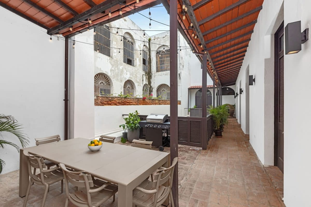 An outdoor terrace is highlighted by a spacious dining table surrounded by six chairs, with a barbecue grill positioned nearby. The area is bordered by white walls and adorned with potted plants, providing a cozy, inviting space for gathering and dining.
