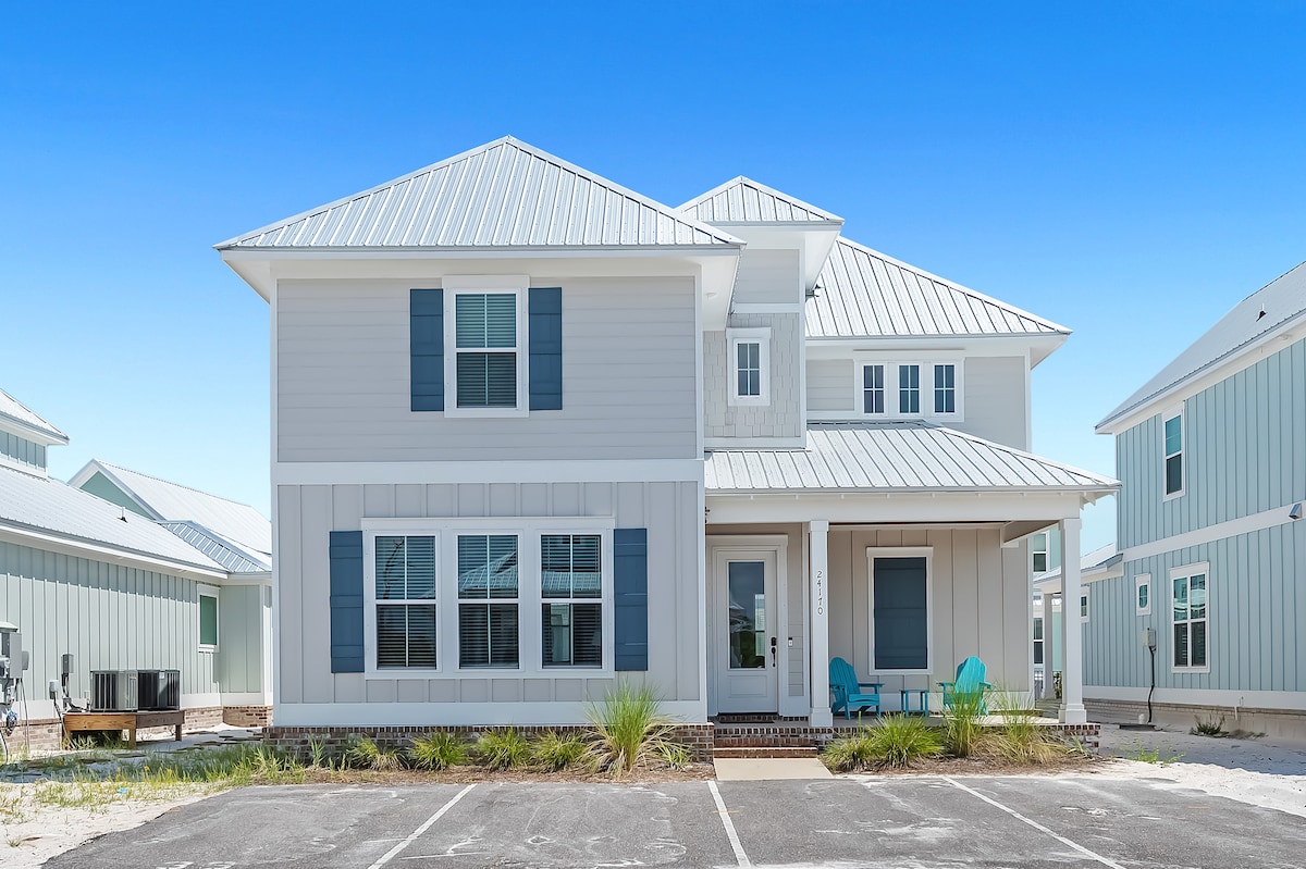 A two-story coastal home features a light-colored exterior with blue shutters. A welcoming front porch includes two chairs and is surrounded by landscaped greenery. The driveway accommodates multiple vehicles, and nearby homes exhibit a similar architectural style under a clear blue sky.