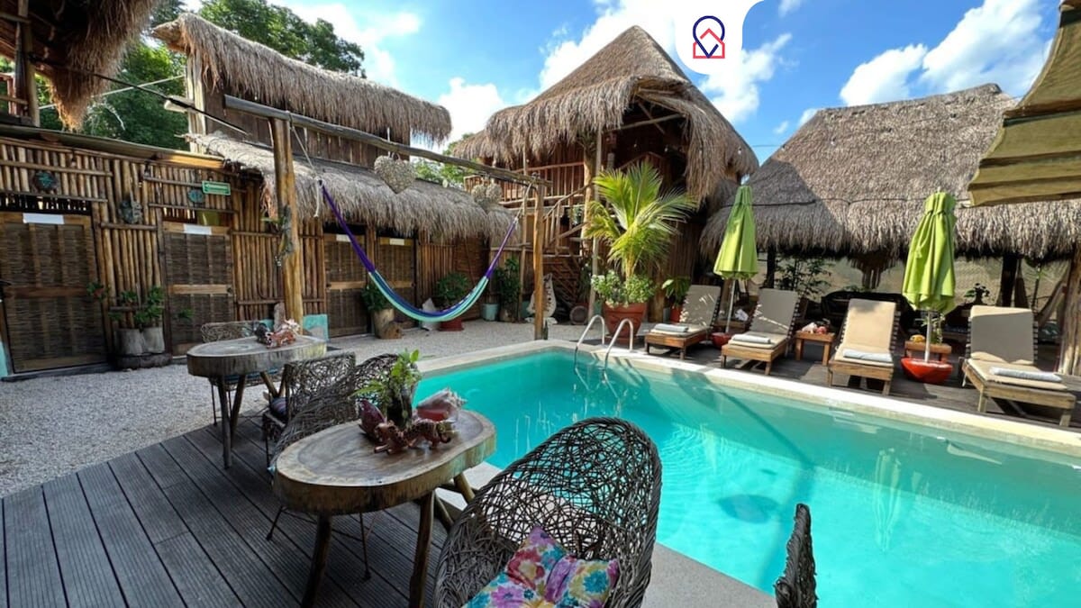 An outdoor relaxation area features a refreshing pool surrounded by sun loungers and shaded tables. Hammocks are hung between thatched structures, while greenery and umbrellas add to the natural ambiance. The deck is made of wooden planks, providing a cozy space for guests to enjoy.