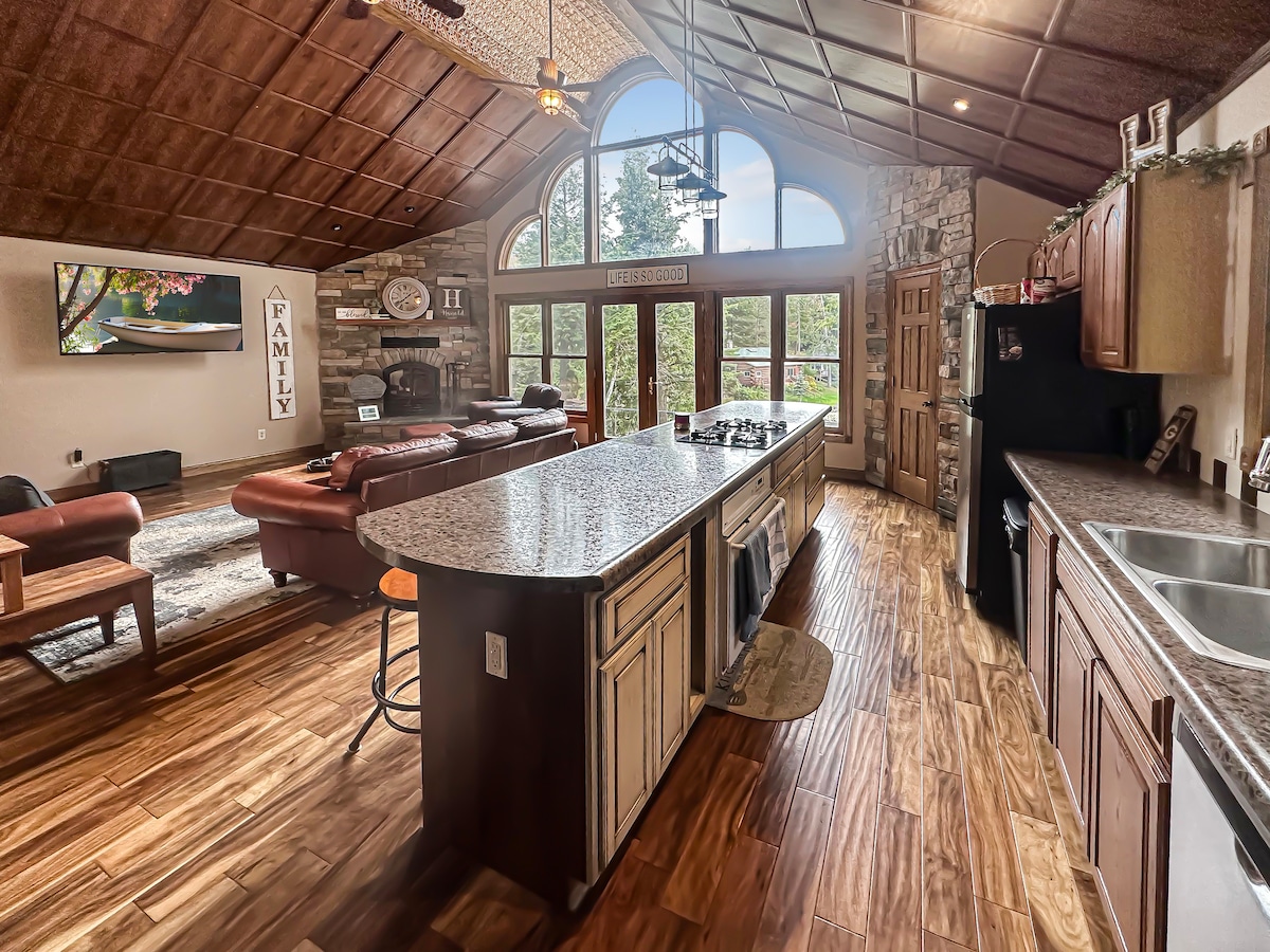 An open-concept living space showcases a large island with a granite countertop in the kitchen area. Natural light floods through tall windows, highlighting a comfortable seating arrangement and a rustic stone fireplace. Wood accents and warm tones create a welcoming atmosphere.