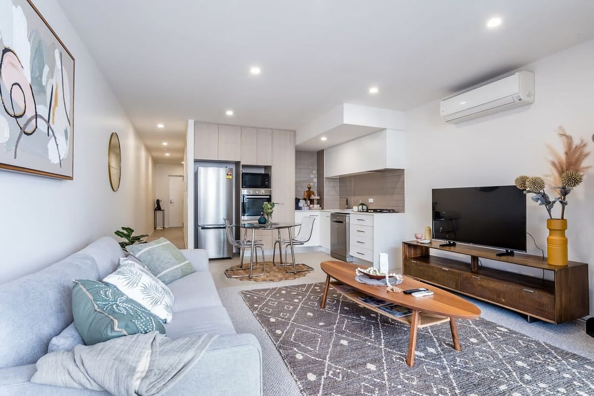 A light-filled living space is presented with a comfortable couch, accent pillows, and a stylish coffee table. An open kitchen is equipped with modern appliances, while a dining table for two is positioned nearby. The area is enhanced by warm wood tones and soft textiles.