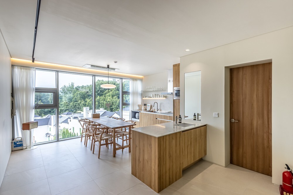 A bright and airy kitchen and dining area features a large island countertop with bar seating. Light wood cabinetry complements the spacious dining table surrounded by wooden chairs. Floor-to-ceiling windows allow natural light to fill the space, with greenery visible outside.