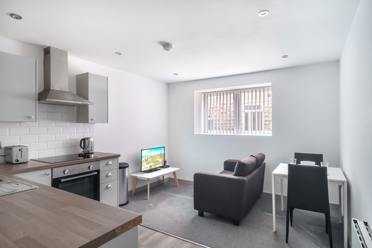 An open plan living area features a snug sofa facing a smart TV. A small dining table with black chairs and a fully equipped kitchen are included. Ample natural light enters through the large window, which is partially covered by vertical blinds.