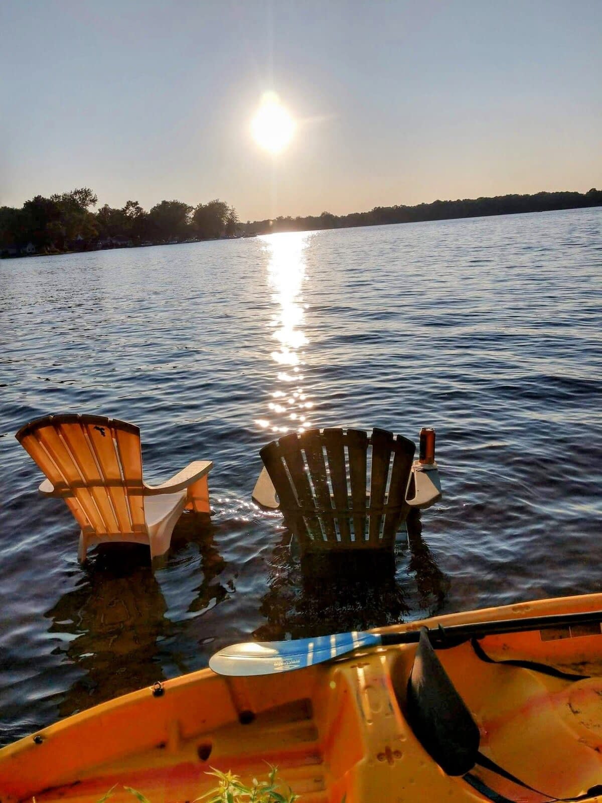 Two adirondack chairs are positioned in shallow water, partially submerged, facing a tranquil lake. The sun sets on the horizon, casting a shimmering reflection across the water's surface. A vibrant kayak rests on the lakeside, adding a touch of color to the serene scene.