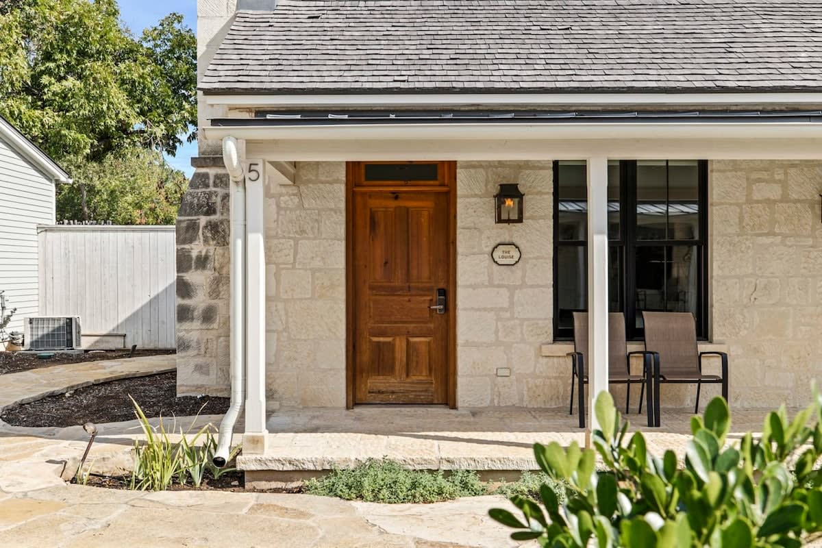 The entrance of the cottage features a solid wooden door surrounded by light-colored stone walls. A welcome sign hangs beside the door. Two modern chairs are placed on the porch, complemented by a small pathway lined with greenery.