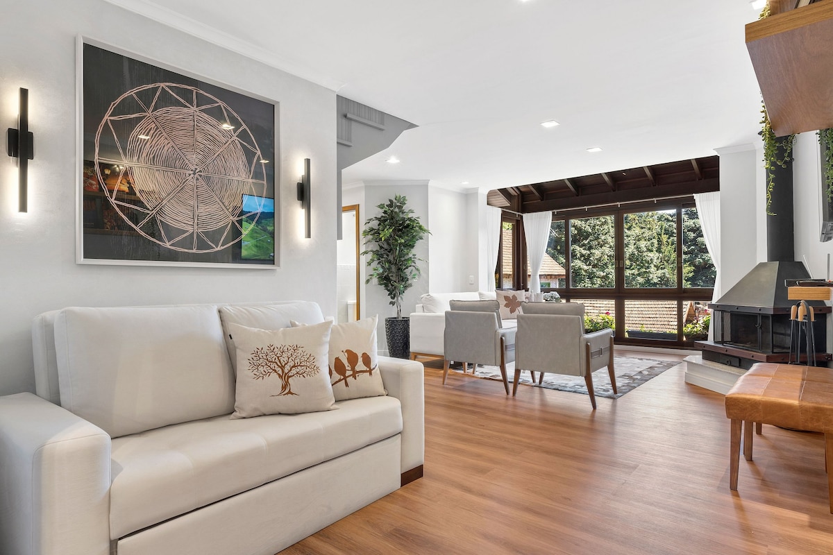 A spacious living area is highlighted by a white sofa with decorative pillows and wooden accents. Large windows allow natural light to fill the space, showcasing green views outside. A modern fireplace is situated next to a wooden bench, adding functionality and style.