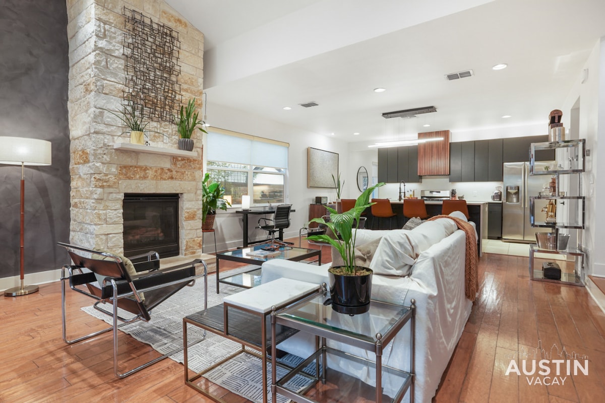 A modern living space is highlighted by a natural stone fireplace and large windows that allow for ample light. A sleek white sofa is complemented by glass tables and a potted plant, with a view of the open-concept kitchen and dining area.
