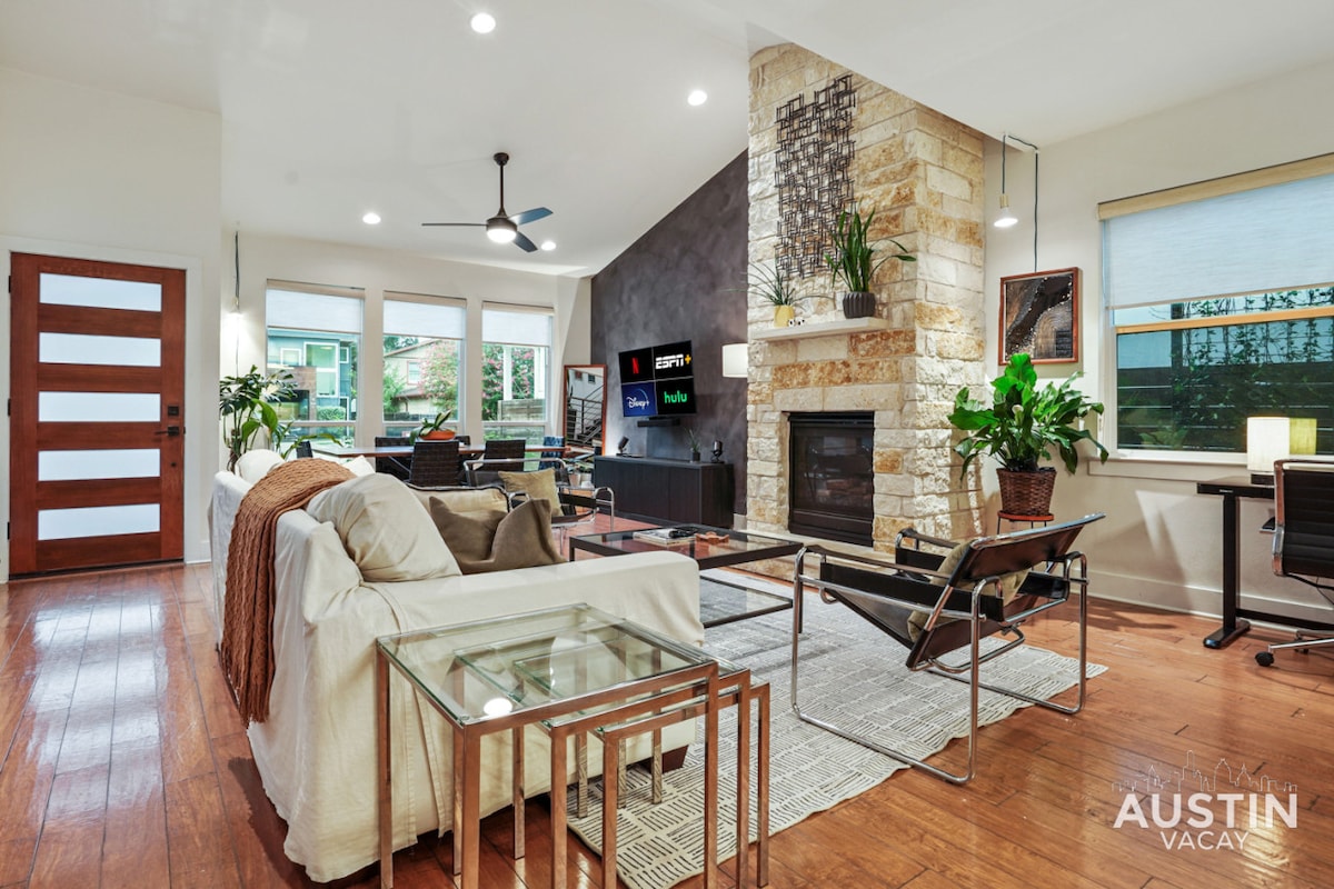 The living room features a mix of textures and colors with a white sofa, sleek glass coffee table, and dark seating. A stone fireplace serves as a focal point, while indoor plants and large windows provide natural light and a connection to the outdoors.