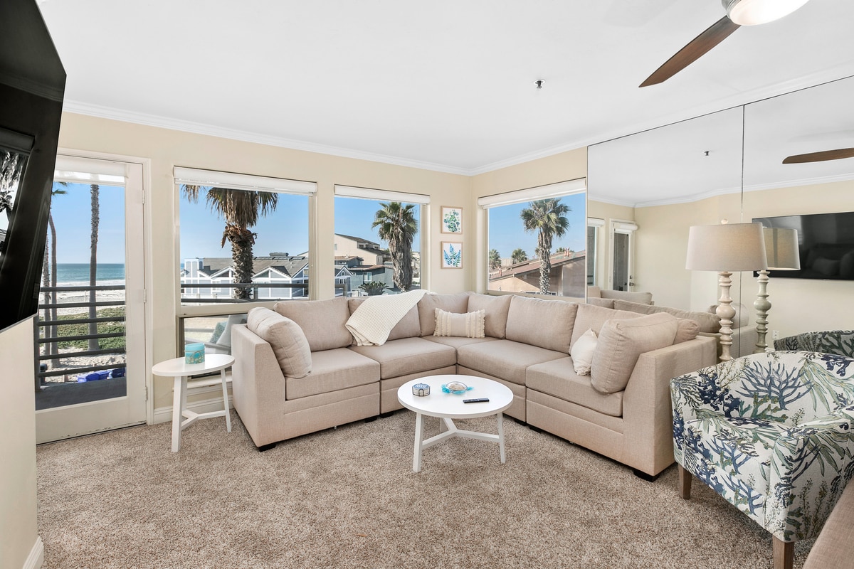 A comfortable living area features a spacious sectional sofa with neutral upholstery, complemented by a patterned armchair. Large windows frame the view of palm trees and the ocean, while a light coffee table holds decorative items. Soft carpet enhances the inviting atmosphere.