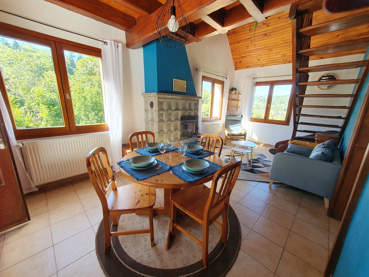 A dining area is furnished with a round wooden table surrounded by four chairs, set against a backdrop of large windows offering scenic views. A blue accent wall complements a cozy seating area and a decorative wood stove adds character to the space.