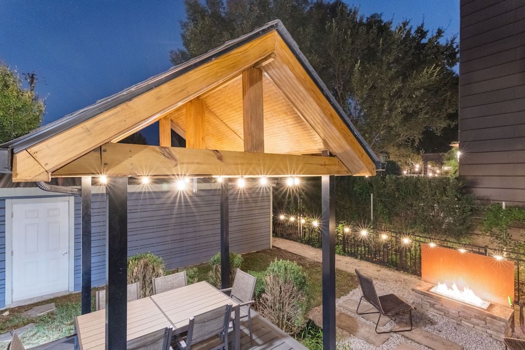 A covered wooden deck features a large dining table surrounded by several chairs. Soft lighting hangs from the structure, providing illumination for evening gatherings. A fire pit is visible nearby, set within a gravel area that enhances the outdoor ambiance.