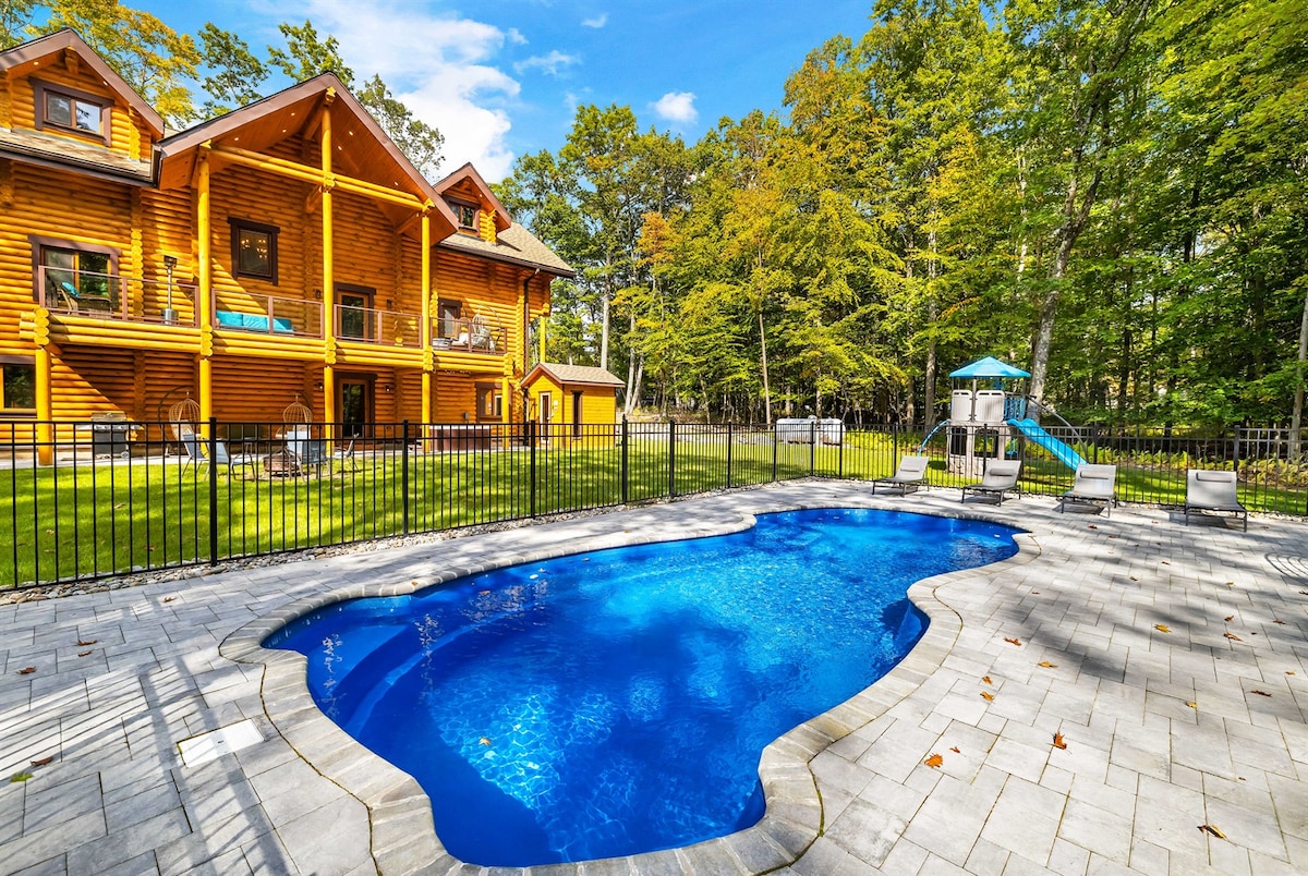 A luxurious log cabin is set amidst lush greenery, featuring an inviting in-ground pool framed by elegant stone pavers. The cabin showcases large windows and multiple decks, providing ample outdoor space for relaxation, while a playground stands nearby, enhancing the family-friendly environment.