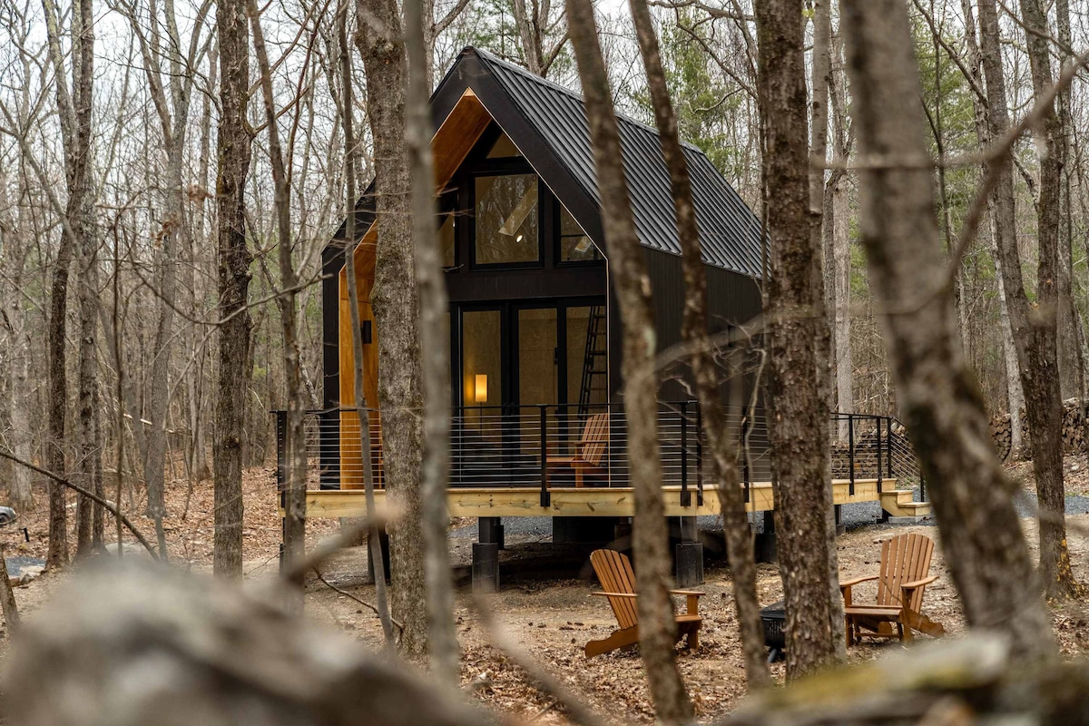 An a-frame cabin is nestled among trees, featuring a modern design with a black exterior and large windows. An L-shaped deck extends from the structure, and several outdoor chairs are positioned nearby, inviting relaxation in the forested setting.