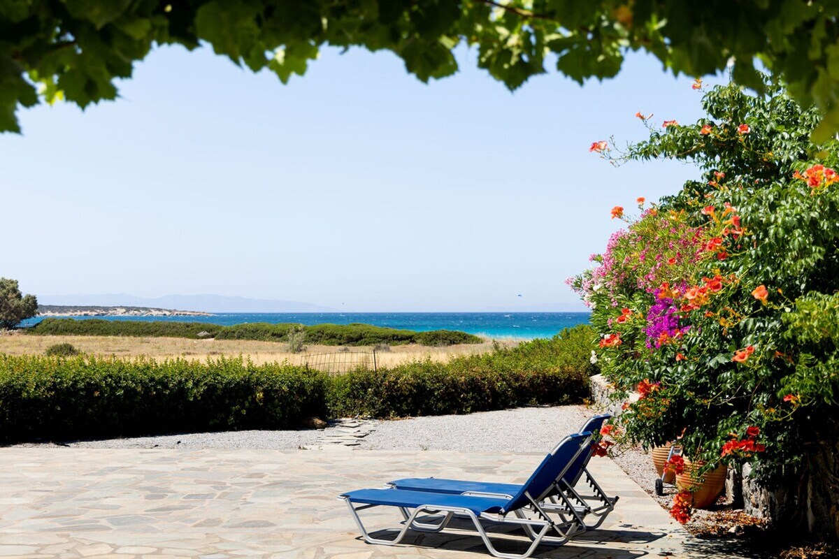 The outdoor terrace features two blue lounge chairs positioned on a stone surface, facing a calm sea. Vibrant flowers in shades of pink and orange frame the scene, while a lush green landscape stretches out towards the horizon, completing the tranquil setting.