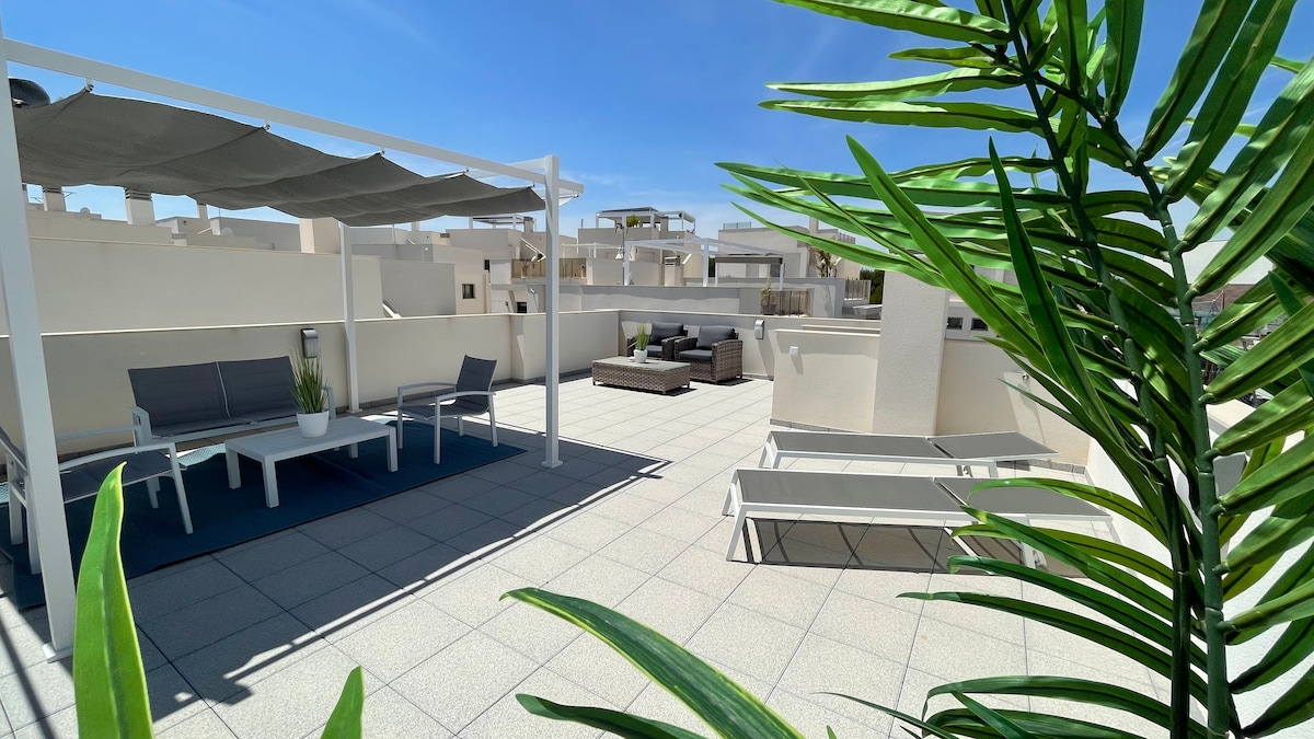 The expansive rooftop solarium features comfortable seating areas and shaded lounges, surrounded by potted greenery. Clear blue skies and sunlight create an inviting atmosphere, while views of the surrounding structures can be seen in the background. This area is ideal for relaxation and enjoying nature.