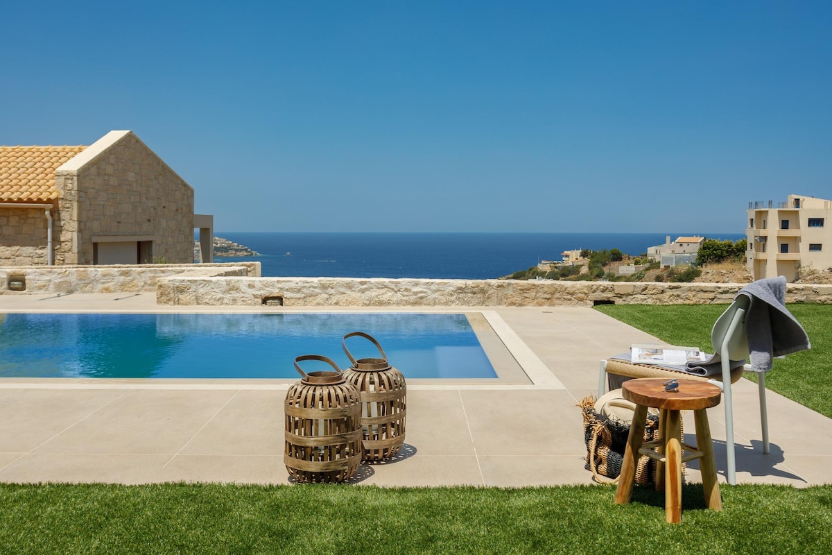 An outdoor sitting area is featured beside a private pool, with clear views of the sea in the background. Two woven stools and a small wooden table are arranged on the patio, surrounded by well-maintained grass. A cozy blanket is neatly draped over a nearby chair.
