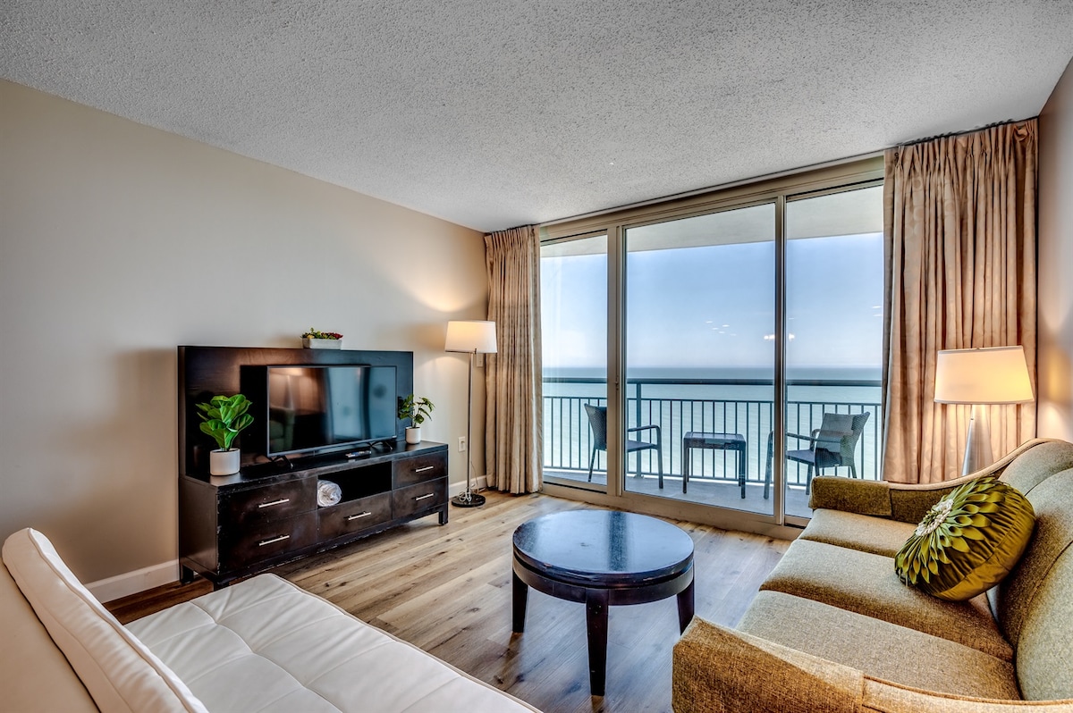 A living area features a comfortable sofa and an accent chair, with a coffee table positioned in the center. A television is mounted on a dark wood console. Floor-to-ceiling windows offer views of the ocean and access to a balcony, framed by light-colored curtains.
