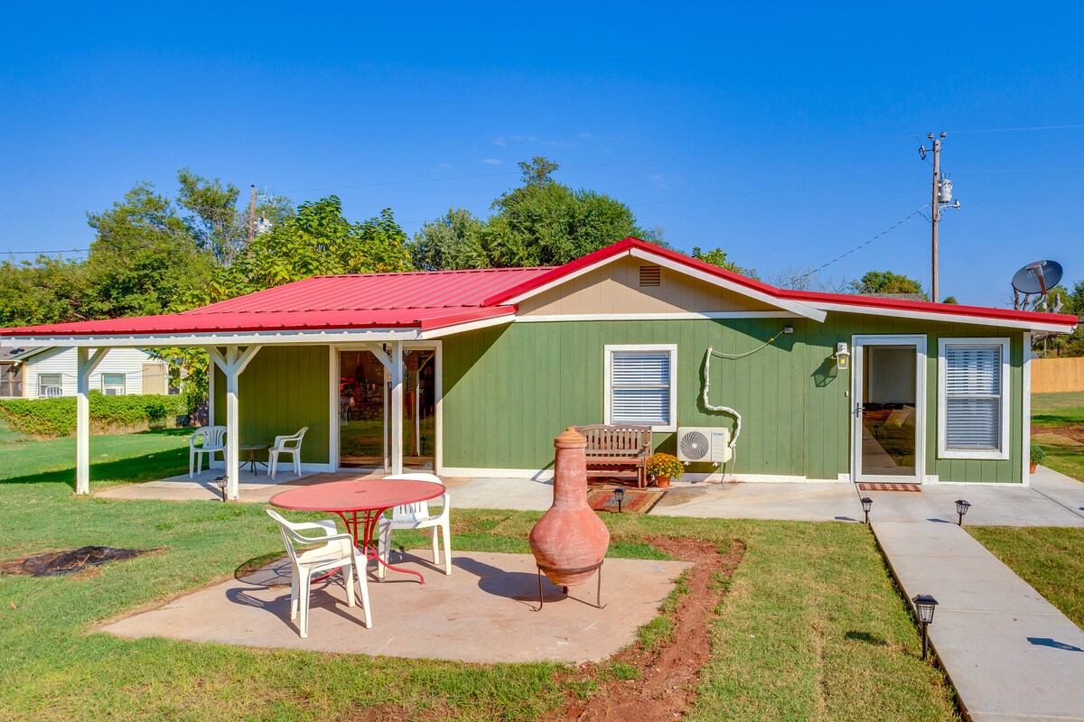 A single-story house with a green exterior and red metal roof is surrounded by a well-kept lawn. A patio area features a round table and chairs, while a clay chiminea stands nearby. The structure has large windows that provide natural light to the interior.
